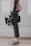 A person is holding a professional video camera with both hands, wearing rolled-up pants, colorful sneakers, and white socks. The background consists of a white brick wall and wooden floor.
