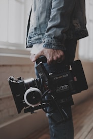 A person holds a professional video camera, wearing a denim jacket. The camera appears to be a high-end model, possibly used for filmmaking or professional video production.