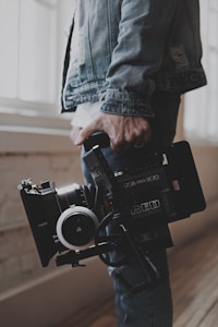 A person holds a professional video camera, wearing a denim jacket. The camera appears to be a high-end model, possibly used for filmmaking or professional video production.