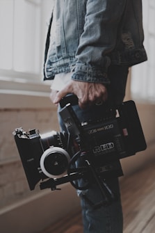 A person holds a professional video camera, wearing a denim jacket. The camera appears to be a high-end model, possibly used for filmmaking or professional video production.