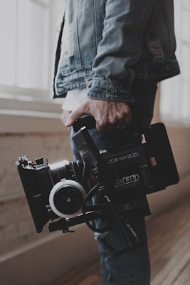 A person holds a professional video camera, wearing a denim jacket. The camera appears to be a high-end model, possibly used for filmmaking or professional video production.