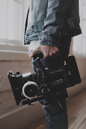 A person holds a professional video camera, wearing a denim jacket. The camera appears to be a high-end model, possibly used for filmmaking or professional video production.