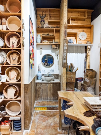 A small, rustic pottery workshop featuring wooden shelves filled with various clay pots and decorative items. A sink basin made of stone is set into a wooden counter, with a round mirror above it. The shelves contain ceramic molds, masks, and colorful fabric displays. In the corner are storage boxes and art supplies.