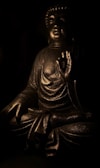 A bronze-colored statue of Buddha is depicted with a serene expression, seated in a meditative pose with one hand raised in a gesture of peace and enlightenment. The statue is illuminated by gentle, low lighting that highlights its intricate details and textured surface, creating a tranquil and reflective ambiance.