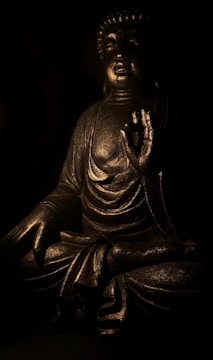 A bronze-colored statue of Buddha is depicted with a serene expression, seated in a meditative pose with one hand raised in a gesture of peace and enlightenment. The statue is illuminated by gentle, low lighting that highlights its intricate details and textured surface, creating a tranquil and reflective ambiance.