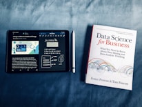 An iPad displaying data visualization content next to a book titled 'Data Science for Business.' The iPad is showing notes and a diagram about dashboard design, accompanied by a stylus placed between the iPad and the book.