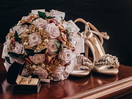 A detailed wedding-themed arrangement featuring a bouquet of artificial roses and pearls placed on a polished wooden surface. Nearby, a pair of elegant high-heeled shoes adorned with gemstones and an open jewelry box containing a ring are visible.