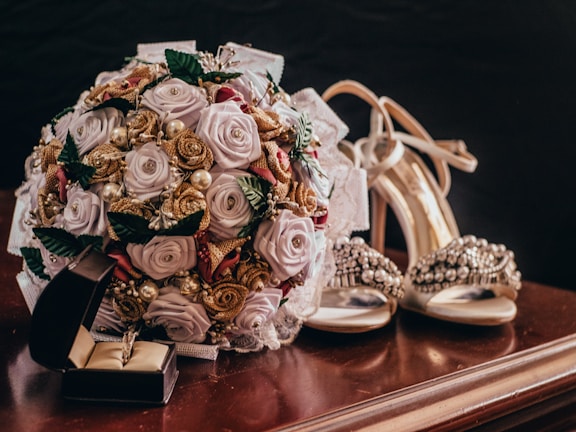 A detailed wedding-themed arrangement featuring a bouquet of artificial roses and pearls placed on a polished wooden surface. Nearby, a pair of elegant high-heeled shoes adorned with gemstones and an open jewelry box containing a ring are visible.