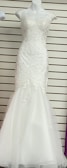 A white, floor-length gown displayed on a mannequin. The gown features intricate lace detailing on the bodice and cap sleeves with a fitted form that flares into a subtle mermaid hem. The background has horizontal lines, possibly from wall paneling or shelves.