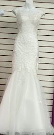 A white, floor-length gown displayed on a mannequin. The gown features intricate lace detailing on the bodice and cap sleeves with a fitted form that flares into a subtle mermaid hem. The background has horizontal lines, possibly from wall paneling or shelves.