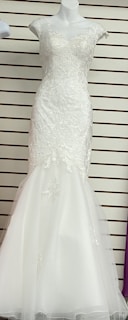A white, floor-length gown displayed on a mannequin. The gown features intricate lace detailing on the bodice and cap sleeves with a fitted form that flares into a subtle mermaid hem. The background has horizontal lines, possibly from wall paneling or shelves.