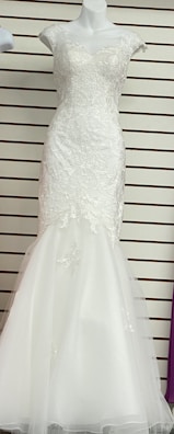 A white, floor-length gown displayed on a mannequin. The gown features intricate lace detailing on the bodice and cap sleeves with a fitted form that flares into a subtle mermaid hem. The background has horizontal lines, possibly from wall paneling or shelves.