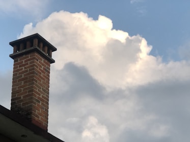 A brick chimney extends upward against a backdrop of large, fluffy white clouds in a clear blue sky. The structure appears to be part of a house roof, visible at the bottom left corner of the image.