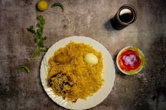 A plate filled with yellow rice, topped with a boiled egg and a potato, garnished with cilantro. Next to the plate, there is a small bowl containing sliced tomatoes, onions, and green chili peppers, forming a colorful salad. A lemon and green chili are placed on the table beside a sprig of cilantro. A dark glass, possibly containing a beverage, is positioned at the top right, creating a balanced composition on a textured, rustic tabletop.