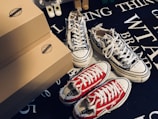 Various pairs of sneakers, including a black and white patterned pair and a red pair with white laces, are positioned on a dark surface with text. Brown shoeboxes are stacked beside the sneakers, and a small toy figure is partially visible in the background.
