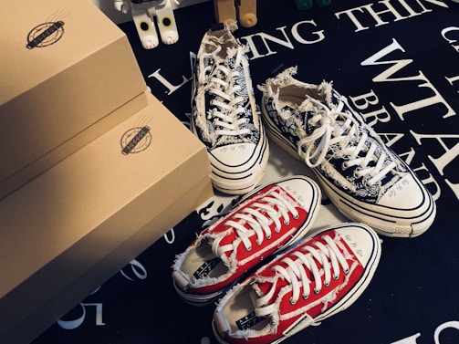 Various pairs of sneakers, including a black and white patterned pair and a red pair with white laces, are positioned on a dark surface with text. Brown shoeboxes are stacked beside the sneakers, and a small toy figure is partially visible in the background.