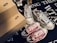 Various pairs of sneakers, including a black and white patterned pair and a red pair with white laces, are positioned on a dark surface with text. Brown shoeboxes are stacked beside the sneakers, and a small toy figure is partially visible in the background.