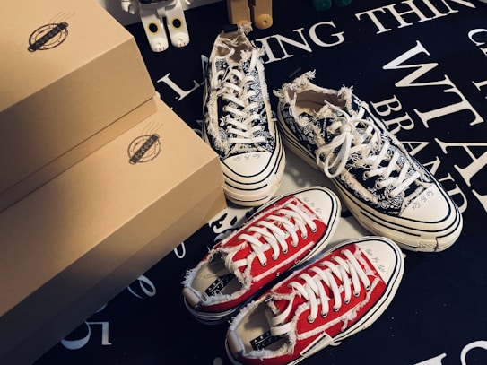 Various pairs of sneakers, including a black and white patterned pair and a red pair with white laces, are positioned on a dark surface with text. Brown shoeboxes are stacked beside the sneakers, and a small toy figure is partially visible in the background.