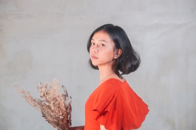 A person with short dark hair is facing sideways, wearing an orange shirt and holding a bunch of dried flowers. The background is a smooth, neutral-colored wall.