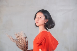 A person with short dark hair is facing sideways, wearing an orange shirt and holding a bunch of dried flowers. The background is a smooth, neutral-colored wall.