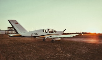A small airplane with the registration 'ESO' is parked on a grassy airfield during sunset. The sky is a mix of soft orange and green hues, and there is a silhouette of a person inside the cockpit. The sun is setting on the horizon, casting a warm glow over the scene.