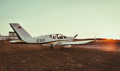A small airplane with the registration 'ESO' is parked on a grassy airfield during sunset. The sky is a mix of soft orange and green hues, and there is a silhouette of a person inside the cockpit. The sun is setting on the horizon, casting a warm glow over the scene.