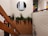 A staircase with wooden railing is adorned with small lights. A planter filled with green foliage is situated on a landing. On the wall above, there is a circular painting of a waterfall. To the right, another colorful painting features abstract designs, including fruit. The walls are a mix of white paint and brick.