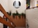 A staircase with wooden railing is adorned with small lights. A planter filled with green foliage is situated on a landing. On the wall above, there is a circular painting of a waterfall. To the right, another colorful painting features abstract designs, including fruit. The walls are a mix of white paint and brick.
