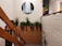 A staircase with wooden railing is adorned with small lights. A planter filled with green foliage is situated on a landing. On the wall above, there is a circular painting of a waterfall. To the right, another colorful painting features abstract designs, including fruit. The walls are a mix of white paint and brick.