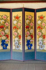 A multi-panel screen with intricate floral designs displayed vertically. Each panel features vibrant patterns of red, white, and yellow flowers with green leaves, set against a yellow background. Blue and grey abstract shapes appear near the bottom of each panel. The panels are bordered in dark blue with a red frame.