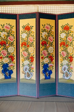 A multi-panel screen with intricate floral designs displayed vertically. Each panel features vibrant patterns of red, white, and yellow flowers with green leaves, set against a yellow background. Blue and grey abstract shapes appear near the bottom of each panel. The panels are bordered in dark blue with a red frame.
