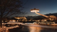 A nighttime scene featuring a futuristic, UFO-like structure hovering above a well-lit park or garden. The area is adorned with numerous twinkling fairy lights, reflecting off the water surface, with dark silhouetted trees in the foreground and background.