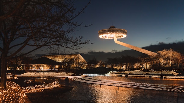 A nighttime scene featuring a futuristic, UFO-like structure hovering above a well-lit park or garden. The area is adorned with numerous twinkling fairy lights, reflecting off the water surface, with dark silhouetted trees in the foreground and background.