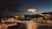 A nighttime scene featuring a futuristic, UFO-like structure hovering above a well-lit park or garden. The area is adorned with numerous twinkling fairy lights, reflecting off the water surface, with dark silhouetted trees in the foreground and background.
