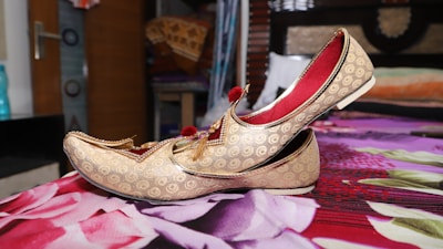 A pair of ornate golden traditional shoes with intricate patterns and red tassels is placed on a floral-patterned bedspread. The shoes have a curved toe and red interior lining. The background includes a partially open wooden door, a water bottle, and fabric items on shelves, suggesting a room setting.