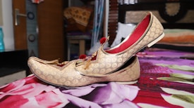 A pair of ornate golden traditional shoes with intricate patterns and red tassels is placed on a floral-patterned bedspread. The shoes have a curved toe and red interior lining. The background includes a partially open wooden door, a water bottle, and fabric items on shelves, suggesting a room setting.