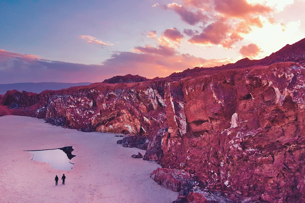 person walking on beach near brown rock formation under cloudy sky during daytime