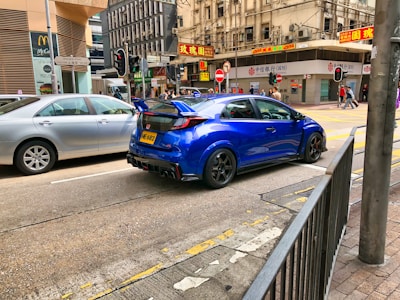 A bright blue sports car with a noticeable rear spoiler is parked on a city street beside a silver sedan. The urban setting features high-rise buildings, a bustling pedestrian area, and various signs written in Chinese. A McDonald's restaurant is visible alongside busy traffic lights and pedestrian crossings.
