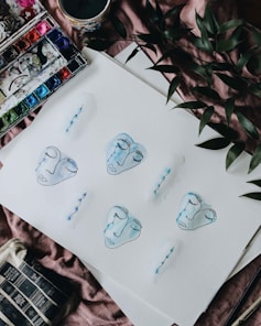 Various watercolor faces with closed eyes and blue outlines are painted on a sheet of paper. The artwork is surrounded by a palette with vibrant colors, a cup with a candle, and green leaves, all resting on a soft, textured surface.