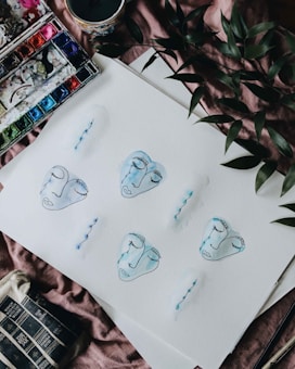 Various watercolor faces with closed eyes and blue outlines are painted on a sheet of paper. The artwork is surrounded by a palette with vibrant colors, a cup with a candle, and green leaves, all resting on a soft, textured surface.
