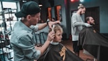 A barber is giving a haircut to a young boy while another man is also receiving a haircut in a well-lit barbershop. The room is equipped with modern furniture and hairdressing tools, and the environment appears relaxed and professional.