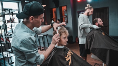 A barber is giving a haircut to a young boy while another man is also receiving a haircut in a well-lit barbershop. The room is equipped with modern furniture and hairdressing tools, and the environment appears relaxed and professional.