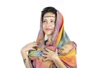 A person wearing a colorful scarf, draped over the head and shoulders, with an intricate pattern and vibrant hues. They are looking slightly upward and have a gentle smile, with hands delicately holding the scarf.