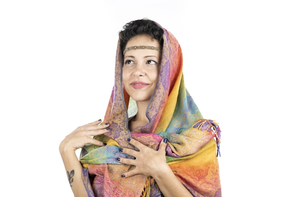 A person wearing a colorful scarf, draped over the head and shoulders, with an intricate pattern and vibrant hues. They are looking slightly upward and have a gentle smile, with hands delicately holding the scarf.