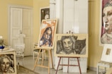 An interior scene features several pieces of art prominently displayed in a room. There are two easels holding expressive, abstract portraits, and additional artwork is visible on the walls and chairs. The setting includes various furniture items such as chairs and a side table, as well as decorative elements like a white dress hanging on the door. The overall ambiance suggests a creative and personal space.