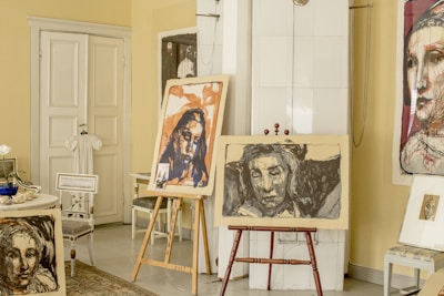 An interior scene features several pieces of art prominently displayed in a room. There are two easels holding expressive, abstract portraits, and additional artwork is visible on the walls and chairs. The setting includes various furniture items such as chairs and a side table, as well as decorative elements like a white dress hanging on the door. The overall ambiance suggests a creative and personal space.