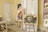 An interior scene features several pieces of art prominently displayed in a room. There are two easels holding expressive, abstract portraits, and additional artwork is visible on the walls and chairs. The setting includes various furniture items such as chairs and a side table, as well as decorative elements like a white dress hanging on the door. The overall ambiance suggests a creative and personal space.