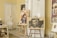 An interior scene features several pieces of art prominently displayed in a room. There are two easels holding expressive, abstract portraits, and additional artwork is visible on the walls and chairs. The setting includes various furniture items such as chairs and a side table, as well as decorative elements like a white dress hanging on the door. The overall ambiance suggests a creative and personal space.