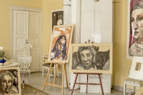 An interior scene features several pieces of art prominently displayed in a room. There are two easels holding expressive, abstract portraits, and additional artwork is visible on the walls and chairs. The setting includes various furniture items such as chairs and a side table, as well as decorative elements like a white dress hanging on the door. The overall ambiance suggests a creative and personal space.