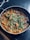 A pan filled with a rich, red-orange curry containing chunks of vegetables or meat, topped with fresh green cilantro leaves. The dish appears freshly cooked and is placed on a dark countertop surface.